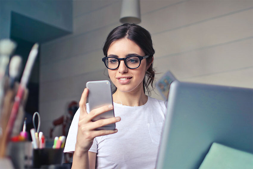 Artsty Woman with Glasses Looking at Cellphone Near Laptop